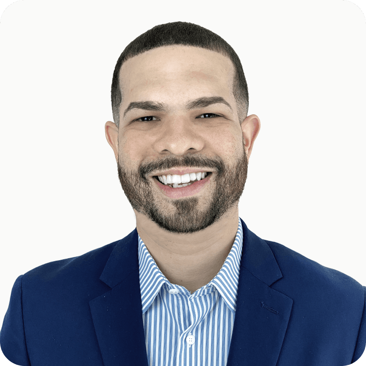 A headshot portrait of a man smiling on a white background in a royal blue suit and a pinstriped dress shirt.